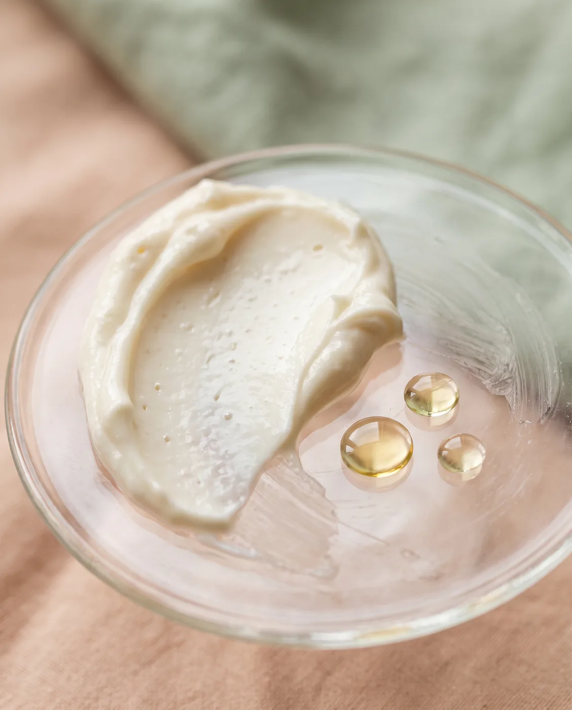 Gros plan d'une crème onctueuse ivoire et de gouttes d'huile dorée sur une plaque de verre, fond pêche et sauge