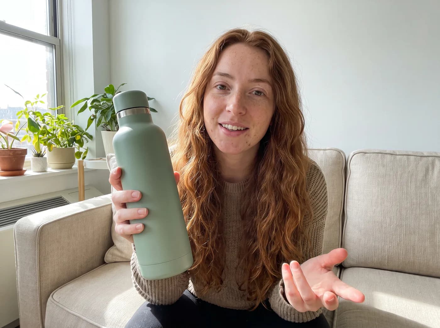 Image de vidéo UGC générée par IA : une femme sur son canapé présente une gourde isotherme vert sauge face caméra