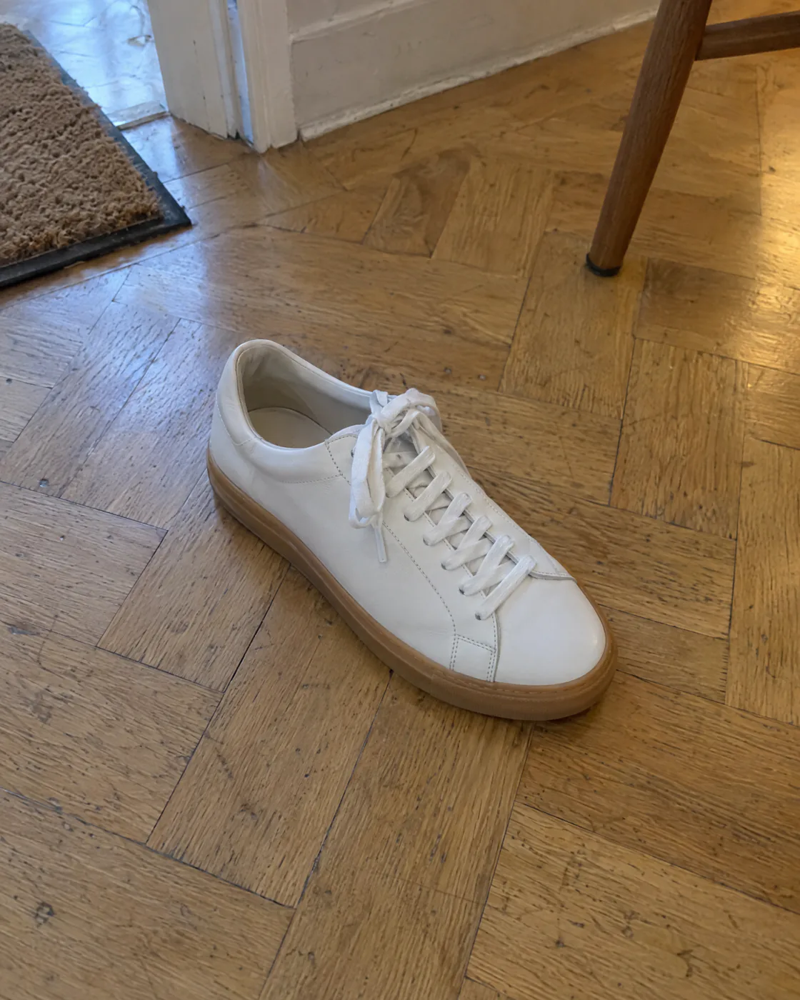 Photo prise au smartphone d'une basket en cuir blanc posée sur un parquet, lumière inégale et décor encombré